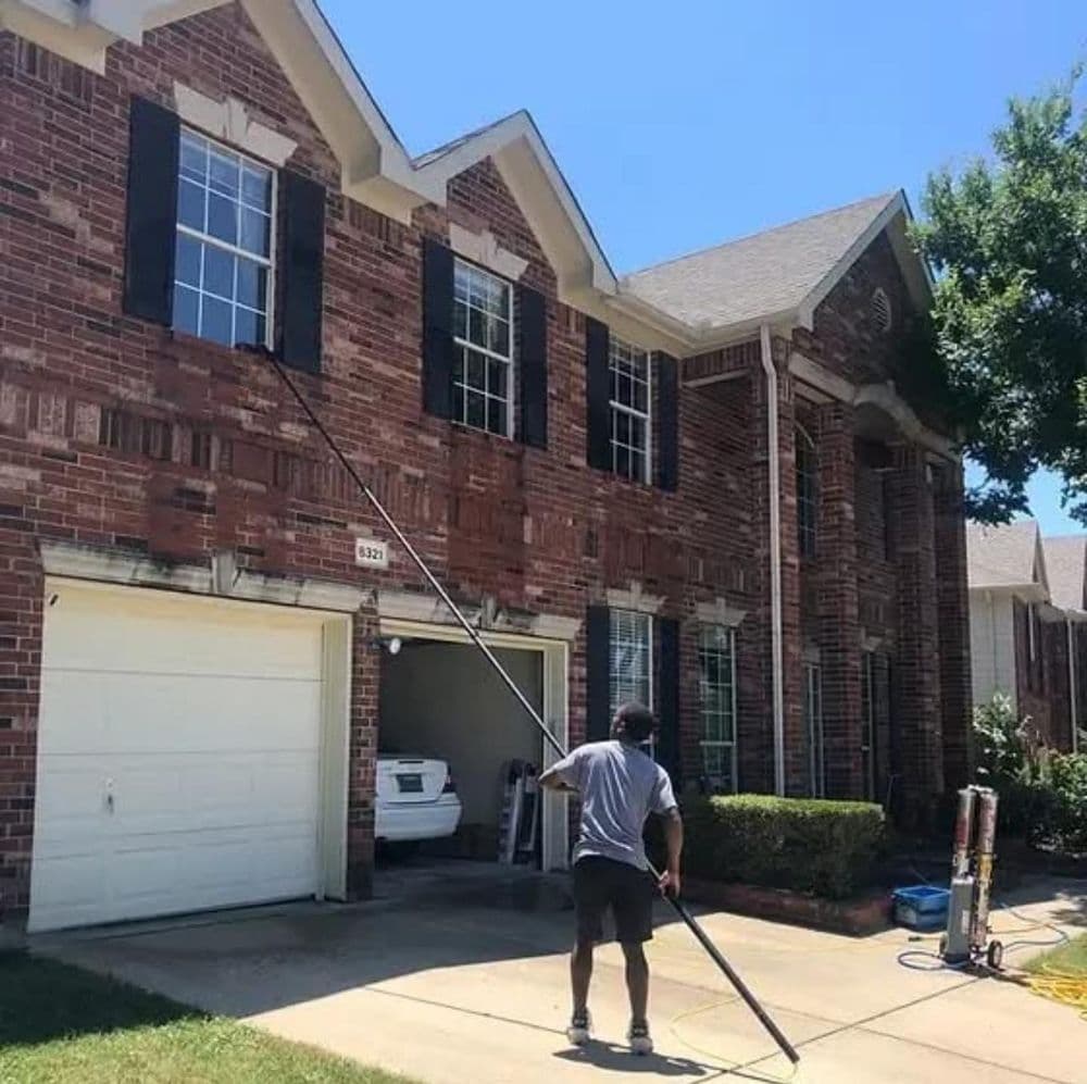 Man using a pole to clean windows on a brick home with a garage in sunny weather.