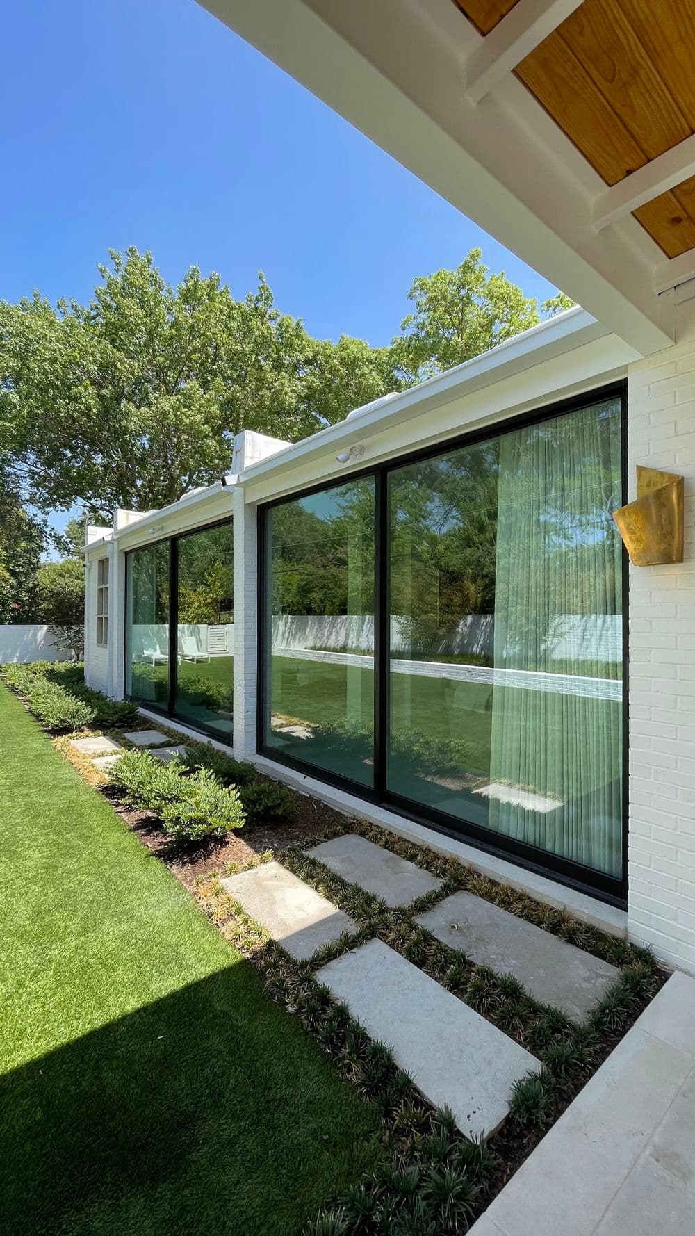 Modern home exterior with large glass windows, manicured lawn, and pathway, under clear blue sky.