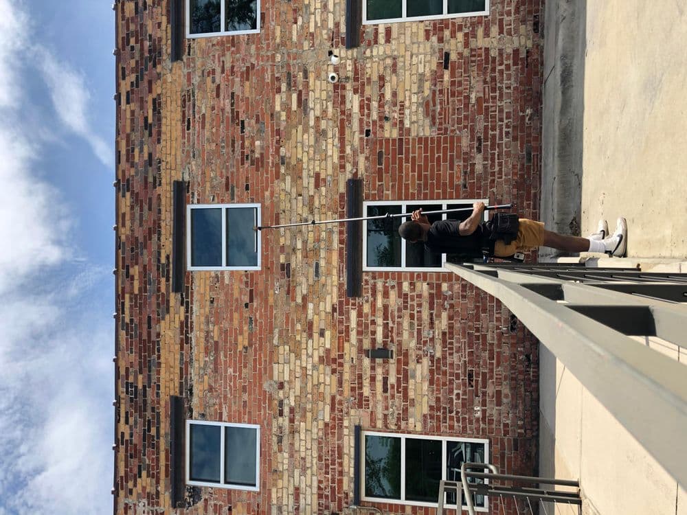 Man cleaning high windows on a brick building with a long pole. Clear blue sky backdrop.