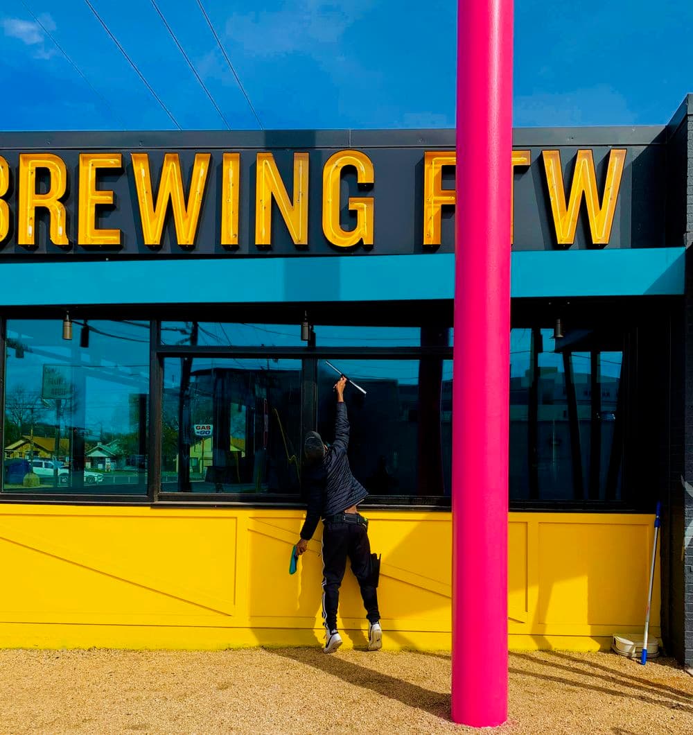 Worker cleaning the window of a brightly colored brewing establishment outdoors.
