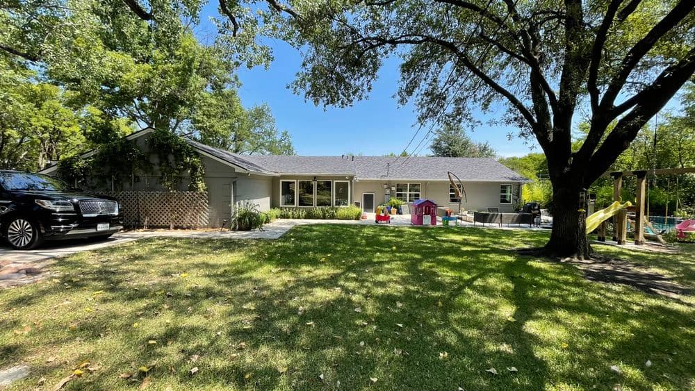 Spacious backyard with playground, trees, and modern home under clear blue sky.