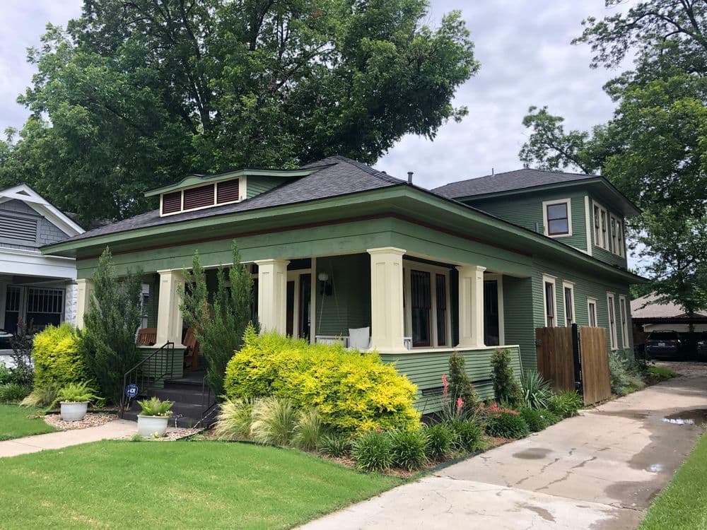 Green two-story house with porch, landscaped front yard, and trees nearby.