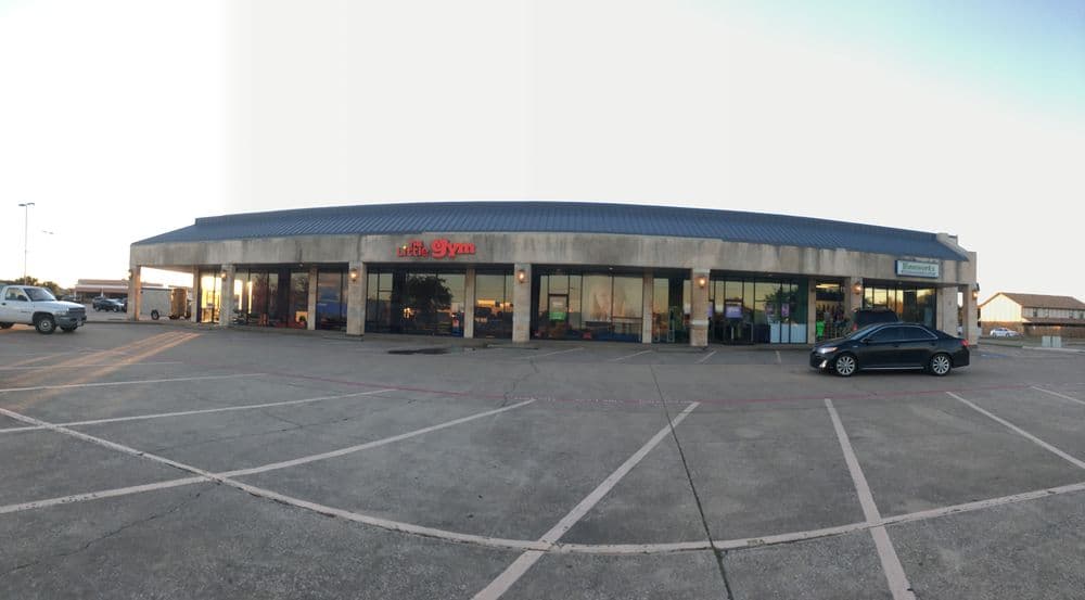 Fitness gym exterior at shopping center with ample parking and clear sky.