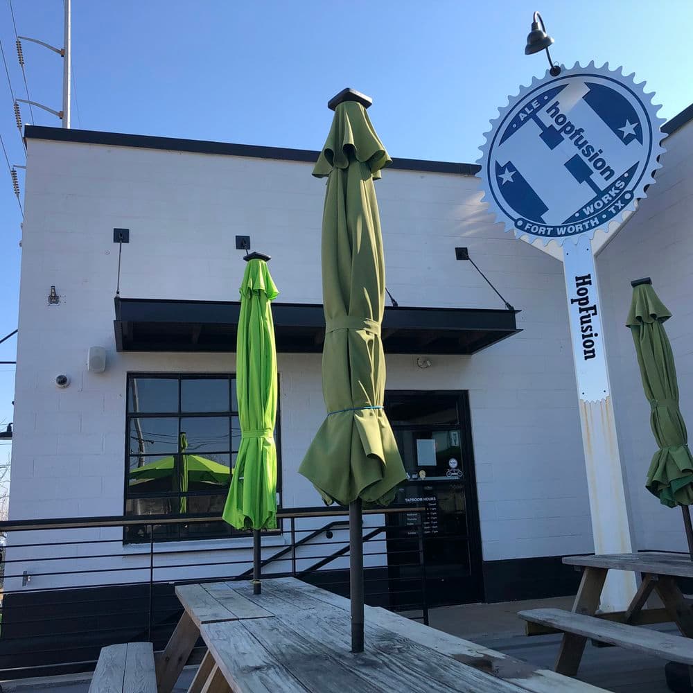 Exterior of Hopfusion brewery in Fort Worth with green umbrellas and picnic tables.